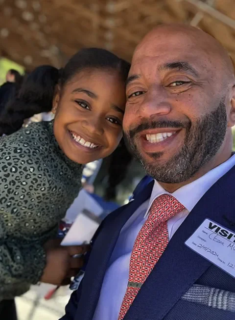 Cezar McKnight smiles in a suit and tie as another person leans in.