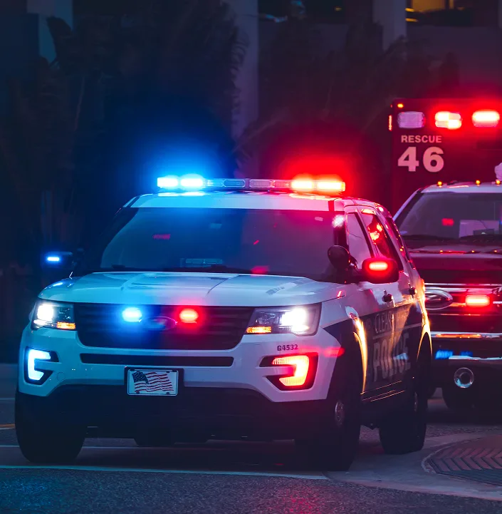 A police SUV with flashing lights drives past at dusk, followed by an emergency vehicle.
