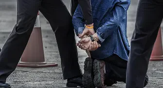 A person in a denim shirt kneels, restrained by two formally dressed individuals.