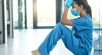 A healthcare worker in blue scrubs sits on the floor, looking stressed.